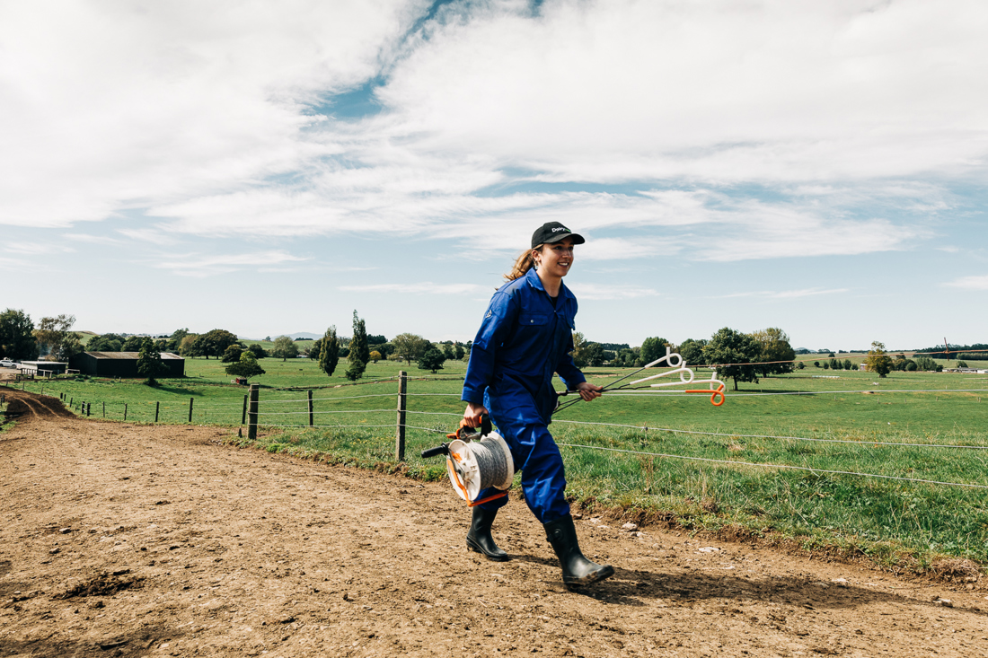 A female farm assistant wearing overalls and gumboots, walking along a farm race carrying fencing stakes.