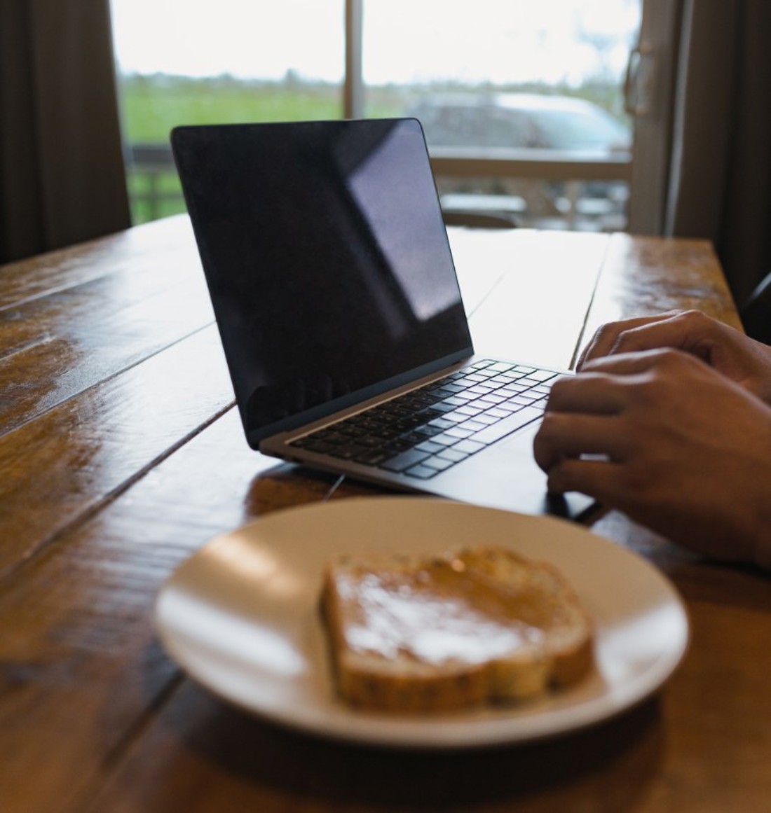 A person using a laptop at a kitchen table with a plate of toast in the foreground
